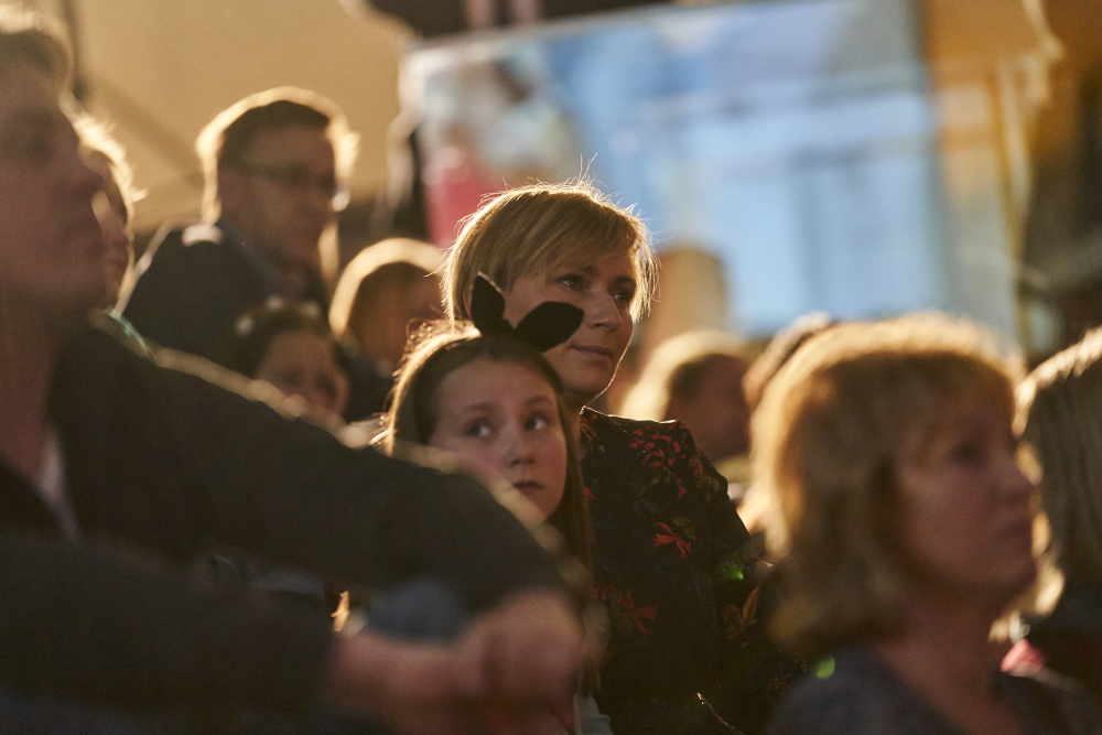 Grupa siedzących ludzi w przytłumionym ciepłym świetle; centralnie kobieta z krótkimi blond włosami siedzi za dziewczynką w opasce z czarnymi uszami, a wokół rozmyte sylwetki innych osób.
