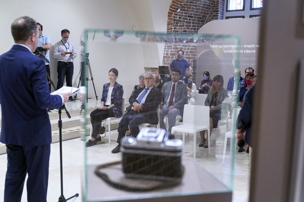 Mężczyzna w granatowym garniturze przemawiający przy mikrofonie po lewej, na pierwszym planie półprzezroczysta szklana gablotka z zabytkowym aparatem, w tle rzędy siedzących osób na białych krzesłach (niektórzy w maseczkach), kamerzyści ze statywami oraz ceglana arkada i fragment projekcji na ścianie.