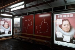 Nocne zdjęcie szklanej wiaty przystanku z podświetlonymi panelami: po bokach dwa plakaty z portretem osoby w kołnierzu typu kryza i przyklejonymi karteczkami, a w środku czerwone panele z białymi ikonami kciuka w dół i kciuka w górę, mokry chodnik na pierwszym planie.