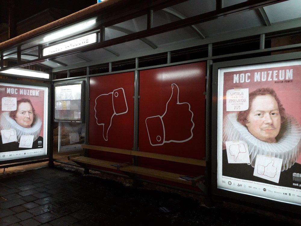 Nocne zdjęcie wiaty przystankowej z drewnianą ławką pośrodku, po bokach podświetlone plakaty z portretem mężczyzny i napisem „MOC MUZEUM”, a między nimi czerwone panele z dużymi białymi szkicami kciuka w górę i w dół; mokry chodnik i śnieg na krawędzi dachu wiaty.