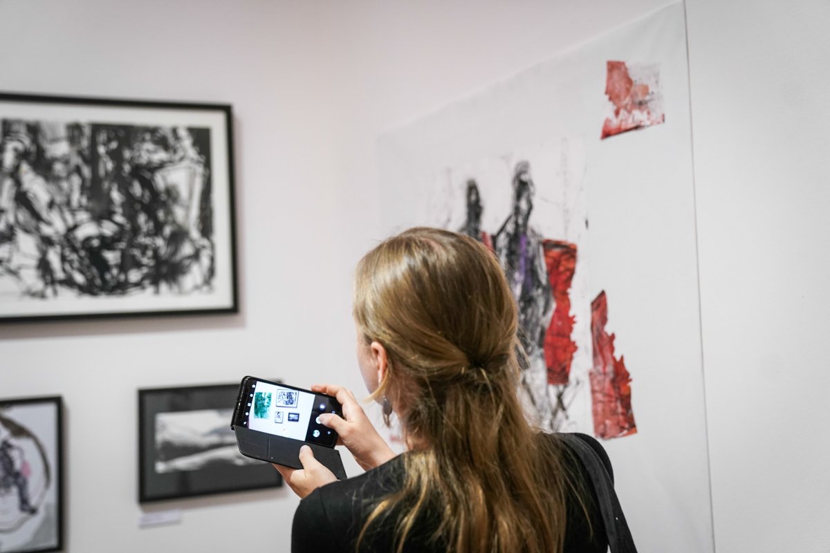 Osoba z długimi jasnobrązowymi włosami spiętymi z tyłu, ubrana na czarno, stoi tyłem w galerii i fotografuje smartfonem w etui oprawione czarno-białe grafiki oraz duży abstrakcyjny obraz z czerwonymi i czarnymi plamami na białej ścianie.