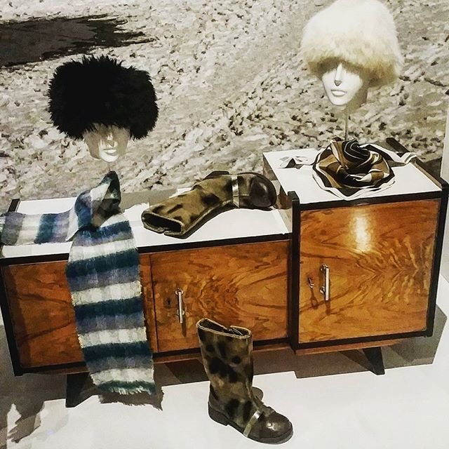 Niska drewniana szafka z dwoma manekinowymi głowami na blacie — lewa w czarnej puszystej czapce, prawa w białej — obok leży pasiasty niebiesko‑szary szalik, rękawice i jedwabna chusta, przed szafką stoi pojedynczy ciemny but w wzór moro, a w tle kamienista powierzchnia.