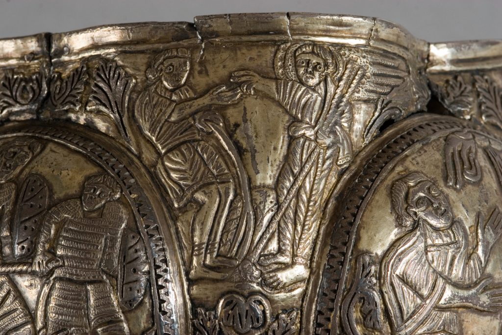 Fragment metalowego reliefu z wyrytymi postaciami w fałdowanych szatach; pośrodku dwie figury z wyciągniętymi rękami, po prawej postać z widocznym skrzydłem, otoczone roślinnymi ornamentami i owalnymi medalionami z mniejszymi wizerunkami, powierzchnia z patyną i drobnymi zarysowaniami.