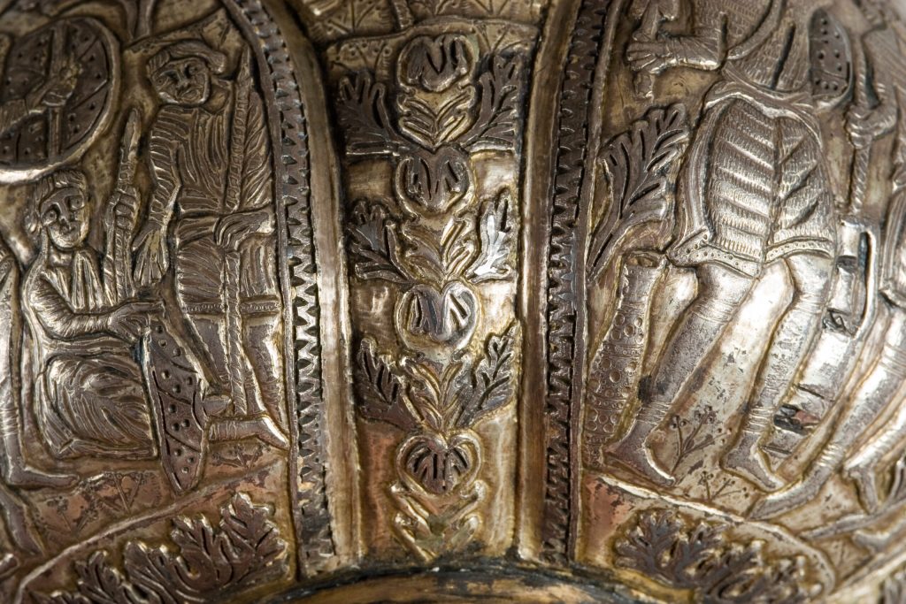 Zbliżenie na metalowy relief o ciemnozłotej patynie z wyżłobionymi stylizowanymi postaciami po bokach, ornamentalnymi pionowymi pasami z motywami liści, ząbkowanymi krawędziami i drobnymi punktowymi zdobieniami.