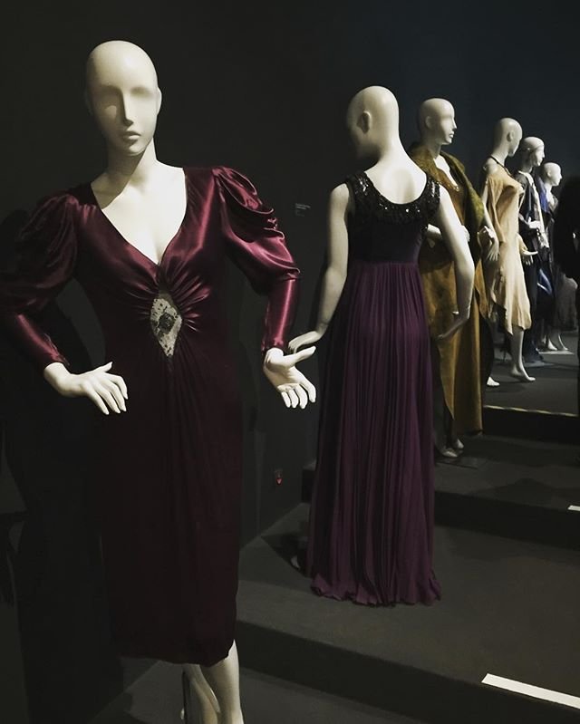 Rząd manekinów w ciemnej galerii ubranych w eleganckie suknie wieczorowe — z przodu bordowa satynowa sukienka z bufkami i ozdobą w talii, za nią długa fioletowa suknia z czarnym zdobionym gorsetem i kolejne manekiny w złotych, beżowych i granatowych kreacjach ustawione na stopniach.