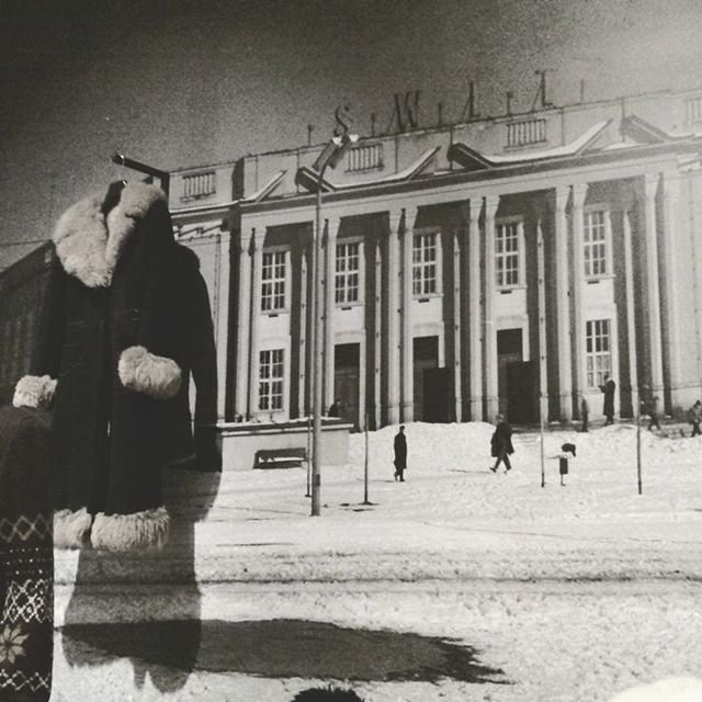 Czarno-białe zdjęcie przedstawiające na pierwszym planie zawieszony płaszcz z futrzanym kołnierzem i mankietami, a w tle rozległy budynek z kolumnami stojący przy zaśnieżonym placu, kilka przechodzących postaci, latarnie i widoczne litery na dachu.