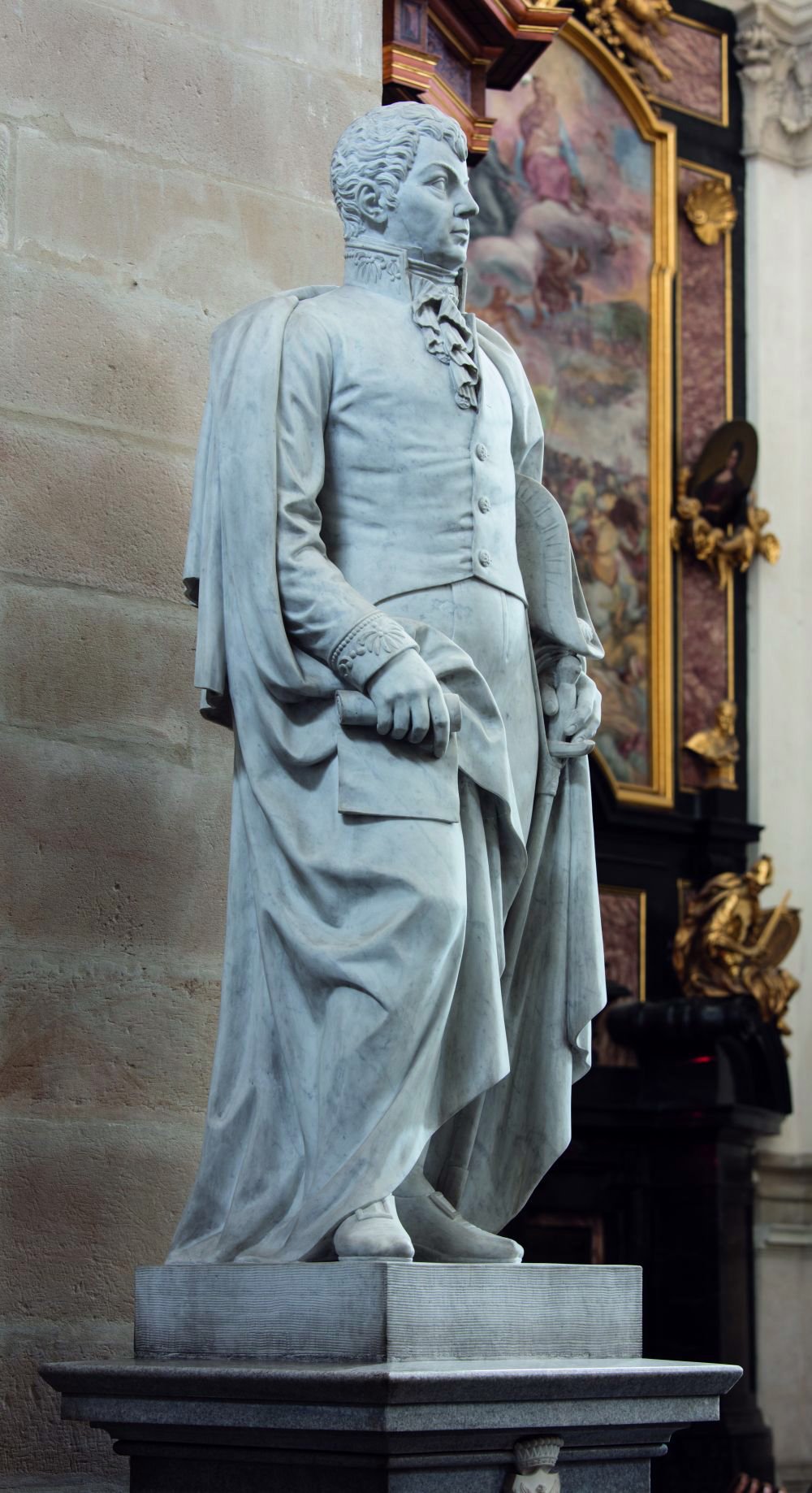 Marmurowa rzeźba stojącej postaci w historycznym stroju i pelerynie, z zrolowanym dokumentem w prawej ręce i dłonią przy pasie, umieszczona na kamiennym cokole przed bogato zdobionym ołtarzem.