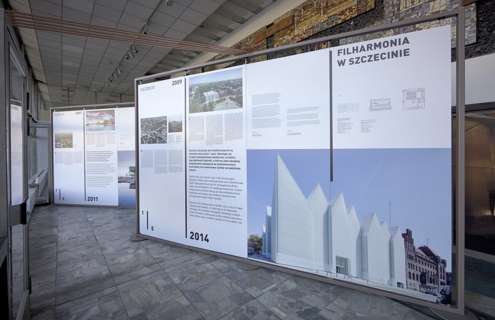 Wnętrze wystawy z dużymi białymi panelami informacyjnymi w metalowych ramach, zawierającymi teksty, zdjęcia i duży render budynku Filharmonii w Szczecinie z charakterystycznym ząbkowanym białym dachem, na marmurowej posadzce pod sufitowymi reflektorami i mozaiką na tylnej ścianie.