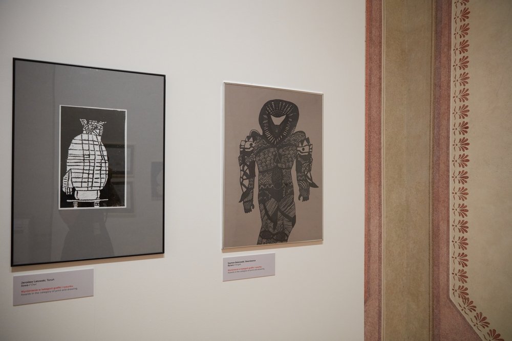 Dwa oprawione czarno-białe grafiki zawieszone obok siebie na jasnej ścianie galerii — po lewej ciemna rama z szarą matą i kontrastową, schematyczną figurą, po prawej jasna ramka z dużą ornamentalnie wzorzystą czarną sylwetką postaci, pod oboma widoczne białe etykiety, a przy prawej krawędzi fragment dekoracyjnego czerwono‑beżowego gzymsu z motywem liści.
