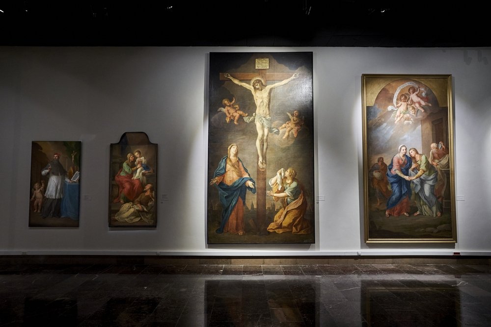 Galeria z białą ścianą i refleksyjną marmurową podłogą, na której wiszą w rzędzie cztery obrazy religijne: od lewej mały portret z dorosłą postacią i dzieckiem, obok wyższy z kobietą trzymającą Dzieciątko w owalnym zwieńczeniu, centralnie duża scena ukrzyżowania z postacią na krzyżu, dwiema klęczącymi kobietami i cherubinami, a po prawej większa rama z grupą postaci i aniołami pod oświetlonym niebem.