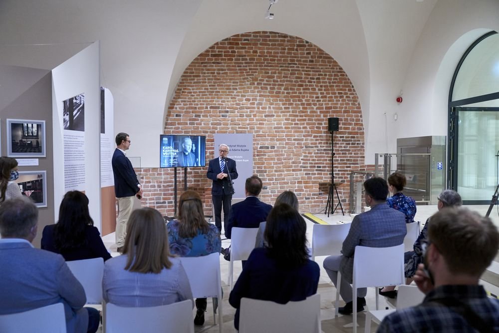 Prezentacja w jasnej sali z dużym ceglanym łukiem w tle, mężczyzna w garniturze przemawia przy mikrofonie przed ekranem z czarno-białym portretem i planszami wystawy, widoczne rzędy białych krzeseł z siedzącą publicznością, stojący mężczyzna po lewej i przeszklone drzwi po prawej.