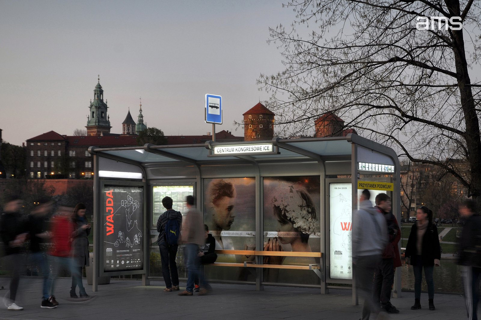 Przystanek z przeszkloną wiatą i znakiem autobusu oraz podświetlonym napisem "CENTRUM KONGRESOWE ICE", wewnątrz plakaty i drewniana ławka, kilka osób stojących i rozmyci przechodnie, a w tle ceglane budynki z wieżami, gołe gałęzie drzewa i zmierzchowe niebo.