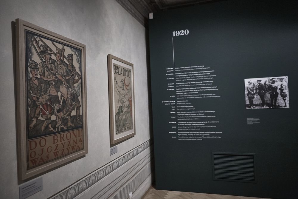 Wnętrze muzealnej sali z jasną ścianą po lewej z dwoma oprawionymi plakatami historycznymi — większym przedstawiającym grupę żołnierzy z widocznym napisem "DO BRONI OJCZYZNA WAS WZYWA" i mniejszym z napisem "BRACIA" — oraz ciemną ścianą po prawej z dużym napisem "1920", pionową linią i kolumną drobnego tekstu oraz małym czarno-białym zdjęciem, nad ozdobną listwą przy podłodze.
