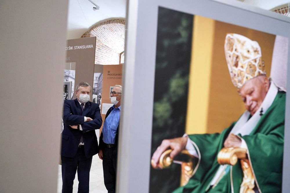 Dwóch mężczyzn w maseczkach (jeden w garniturze ze skrzyżowanymi rękami, drugi w niebieskiej koszuli) stoi w przestrzeni wystawienniczej z panelami informacyjnymi i ceglaną arkadą, po prawej w pierwszym planie duży plakat przedstawiający siedzącego duchownego w zielonych szatach i ozdobnej mitrze.