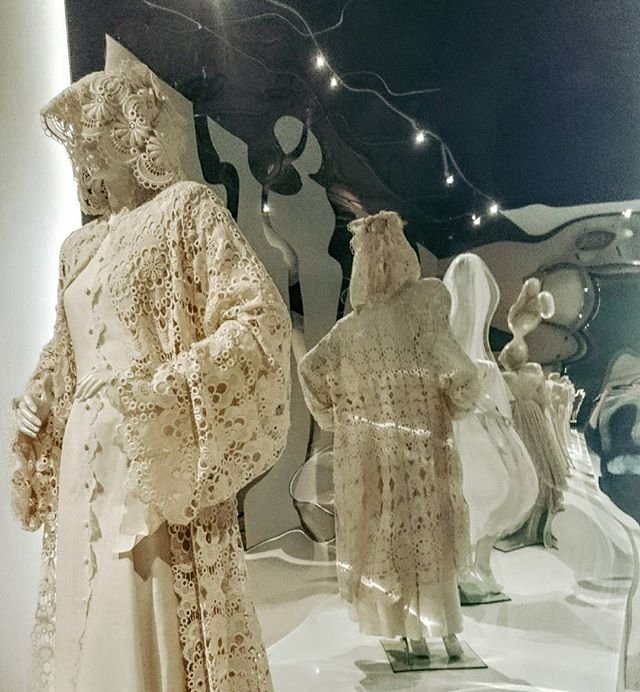 Na pierwszym planie manekin w kremowej, misternie zdobionej koronkowej sukni z kapturem i szerokimi rękawami; w tle odbicia kilku podobnych beżowych koronkowych strojów na falistych lustrzanych panelach oraz migoczące lampki.