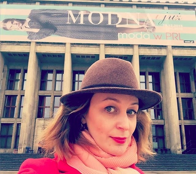 Selfie kobiety w brązowym kapeluszu, różowym szalu i czerwonym płaszczu na tle schodów i wysokich kolumn budynku, nad którym wisi poziomy baner reklamowy z dużym napisem MODNA i sylwetką modelki.