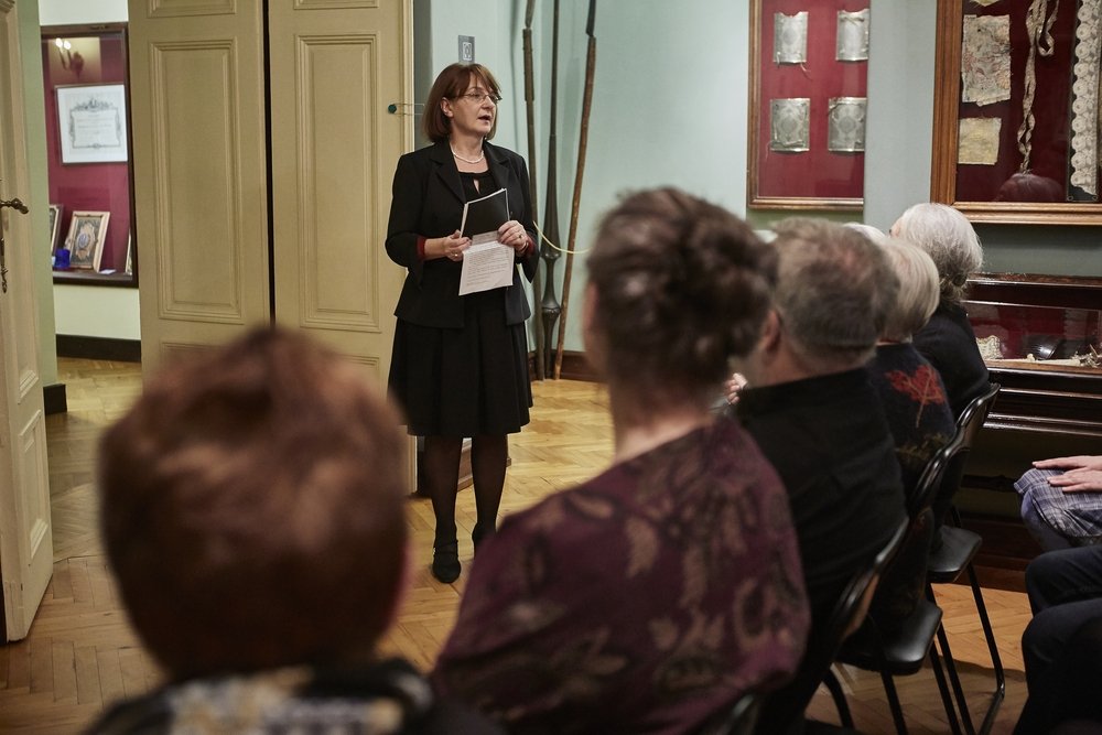 Kobieta w czarnym stroju stoi trzymając kartki przed siedzącą widownią w sali przypominającej muzeum z drewnianą podłogą, gablotami i wiszącymi eksponatami, na pierwszym planie widoczne tylne części głów kilku słuchaczy.