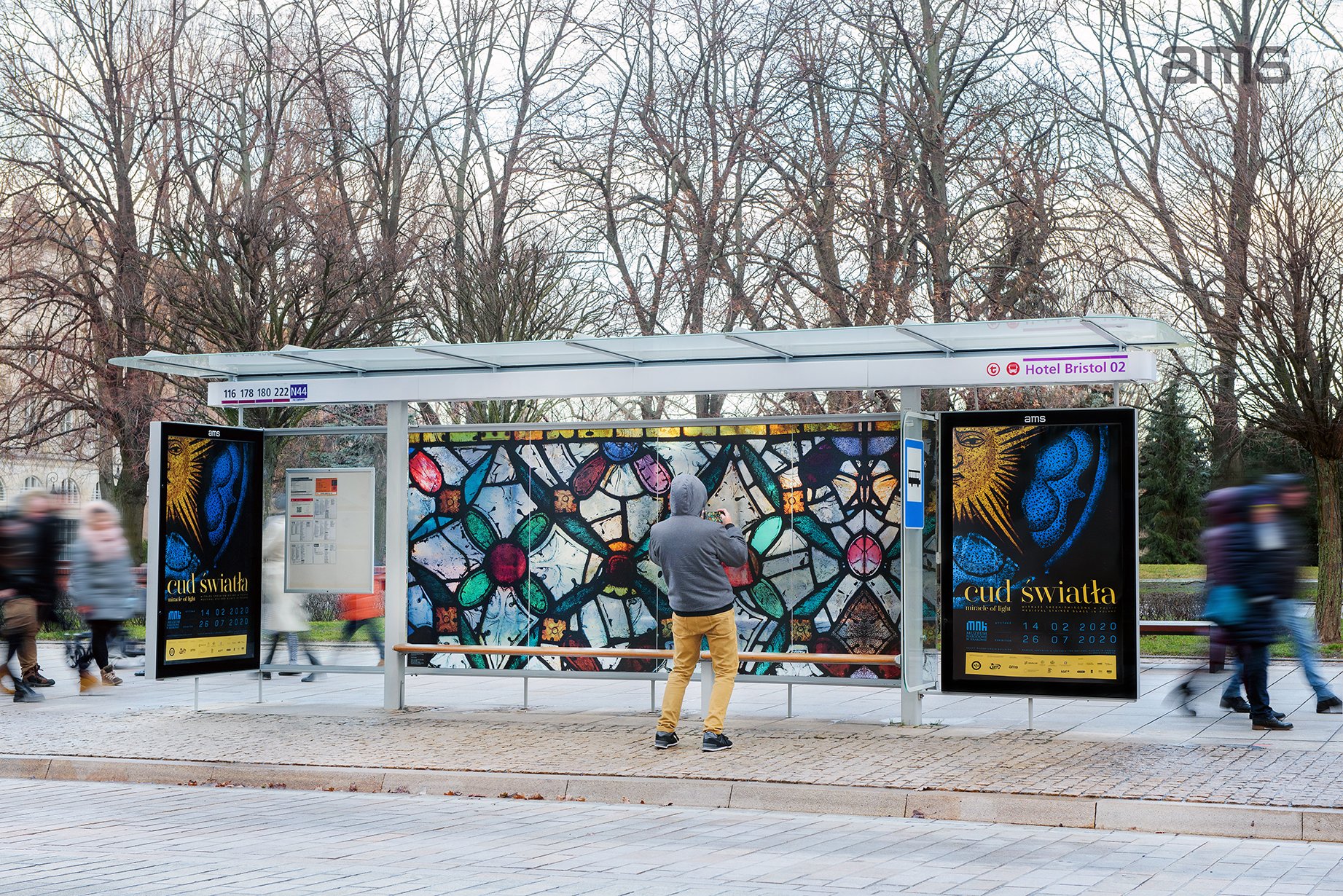 Przystanek autobusowy z przeszklonym dachem i ławką, centralnie osoba w szarej bluzie z kapturem i żółtych spodniach stoi tyłem i fotografuje dużą kolorową panelową grafikę przypominającą witraż; po obu stronach dwa podświetlane plakaty z napisem "cud światła", nad dachem tabliczka z numerami linii i oznaczeniem "Hotel Bristol 02", w tle nagie drzewa i rozmazani przechodnie.