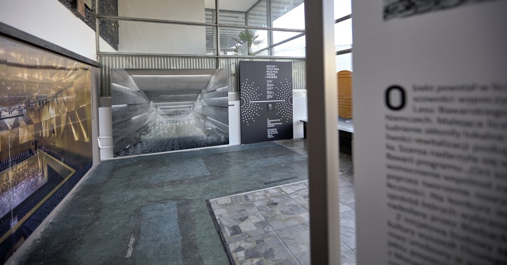 Wnętrze sali wystawowej z dużymi wydrukami architektonicznymi na ścianach: po lewej kolorowy obraz widowni, na środku czarno-biała fotografia betonowej konstrukcji, obok czarna plansza z białym tekstem i punktami, po prawej w pierwszym planie rozmyty panel z tekstem; na podłodze mozaika kamienna i zielonkawy wykładzinowy pas, w tle okna z rośliną w doniczce.
