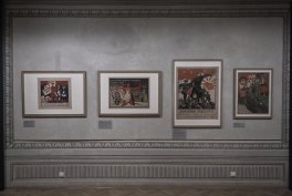 Szara ściana wystawowa z ozdobnymi listwami, na której w jednej linii zawieszono cztery oprawione grafiki/postery różnych rozmiarów z małymi tabliczkami opisowymi pod każdą ramą, nad drewnianą podłogą i listwą przypodłogową.