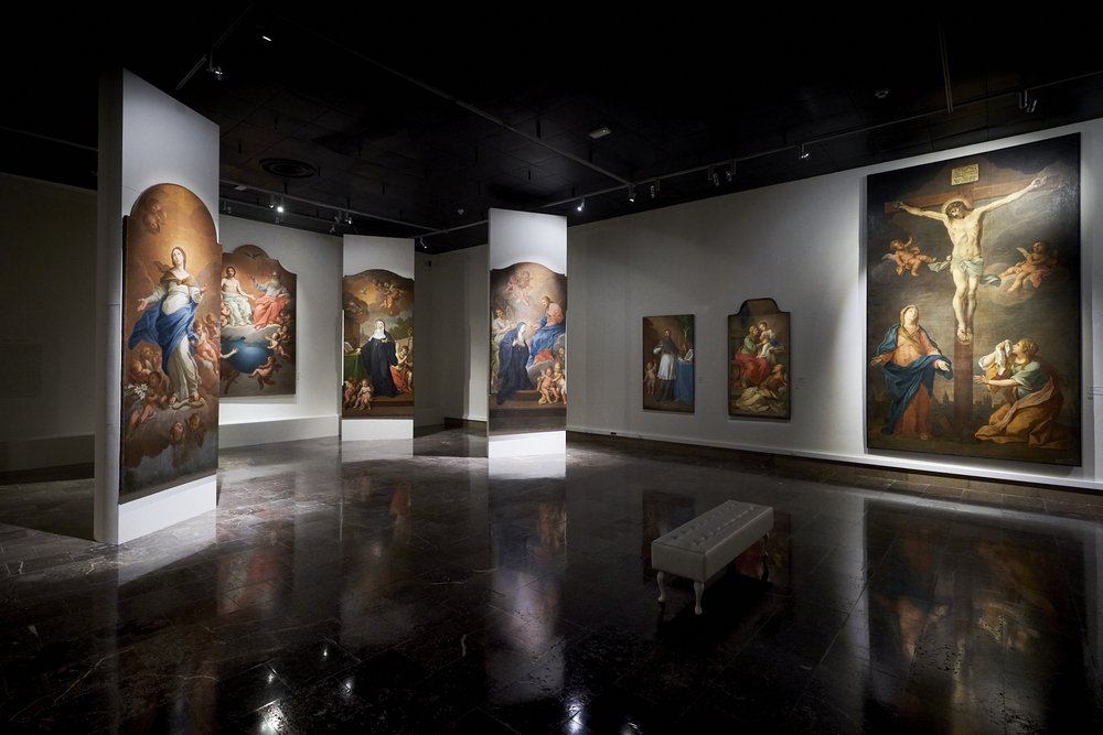 Przestronna, przyciemniona galeria z oświetlonymi, dużymi obrazami o tematyce religijnej zawieszonymi na ścianach i trzech wolnostojących białych panelach, błyszcząca ciemna marmurowa podłoga odbijająca dzieła oraz pojedyncza tapicerowana ławka na środku sali.