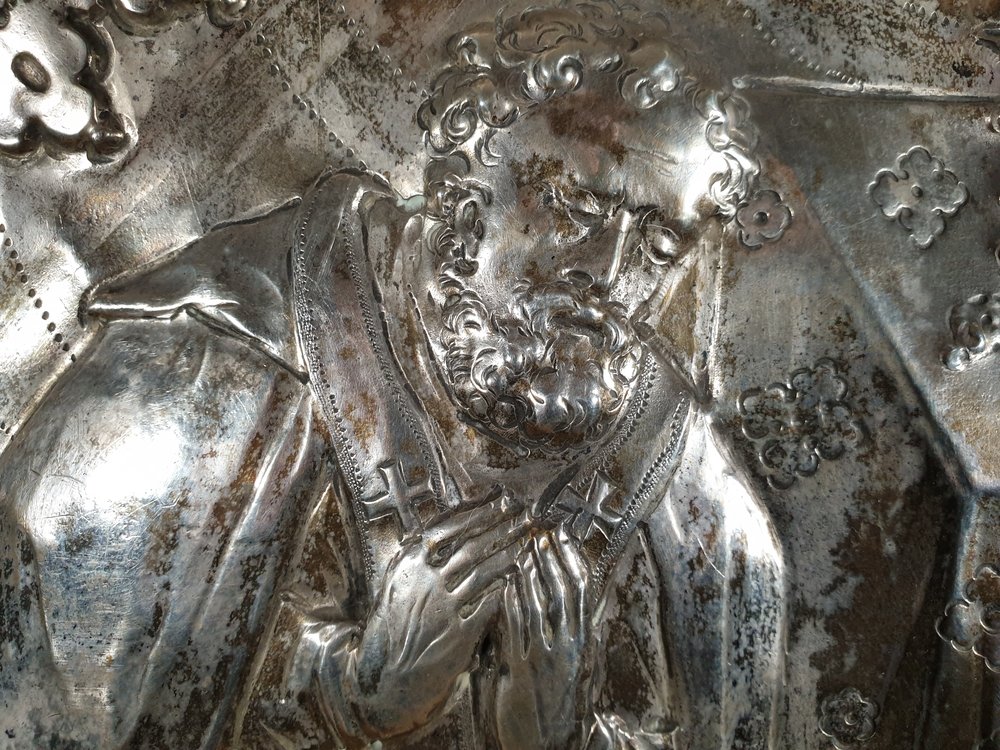 Zbliżenie na srebrny tłoczony relief przedstawiający postać męską z kręconymi włosami i bujną brodą, w liturgicznych szatach z krzyżami, z dłońmi złożonymi przy piersi, na tle kwiatowych ornamentów i miejscowej patyny.