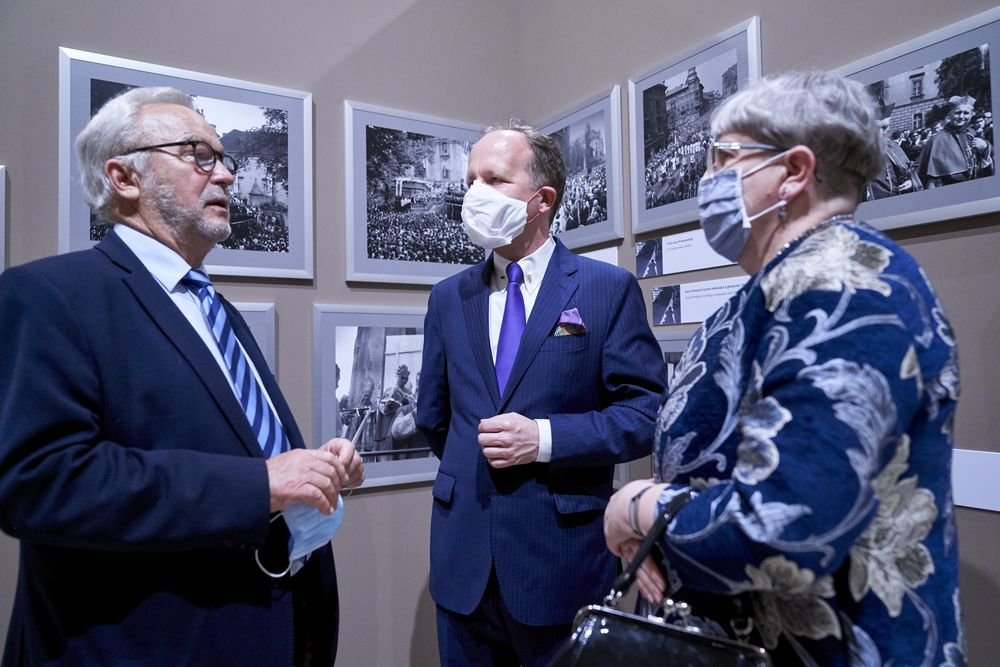 Dwaj mężczyźni i kobieta w medycznych maseczkach stoją przed ścianą z oprawionymi czarno-białymi fotografiami; jeden z mężczyzn trzyma maseczkę w dłoniach, obaj są w garniturach, kobieta ma granatowy wzorzysty płaszcz i czarną torebkę.