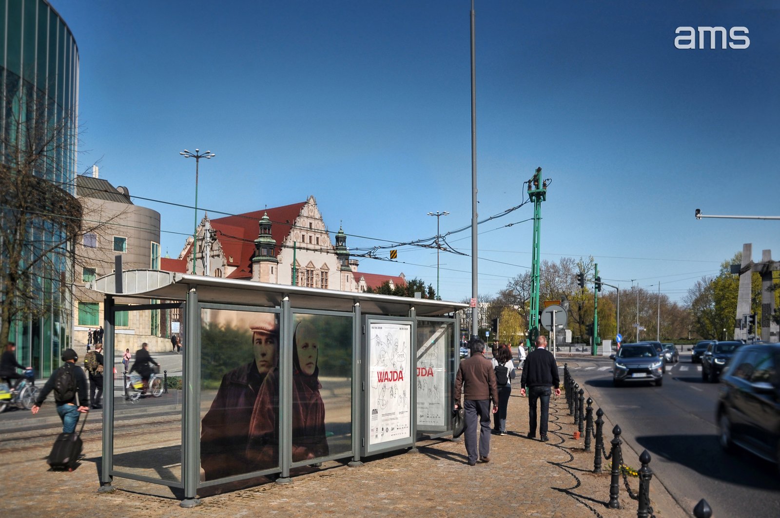 Przystanek tramwajowy z przeszklonymi gablotami i plakatem przedstawiającym dwie osoby na pierwszym planie, obok chodnik z pieszymi i łańcuchem słupków, po prawej wielopasmowa ulica z przejeżdżającymi samochodami, w tle ozdobny zabytkowy budynek z czerwonym dachem oraz nowoczesny szklany gmach, nad ulicą sieć trakcyjna i słupy, a na czystym niebieskim niebie w prawym górnym rogu napis ams.