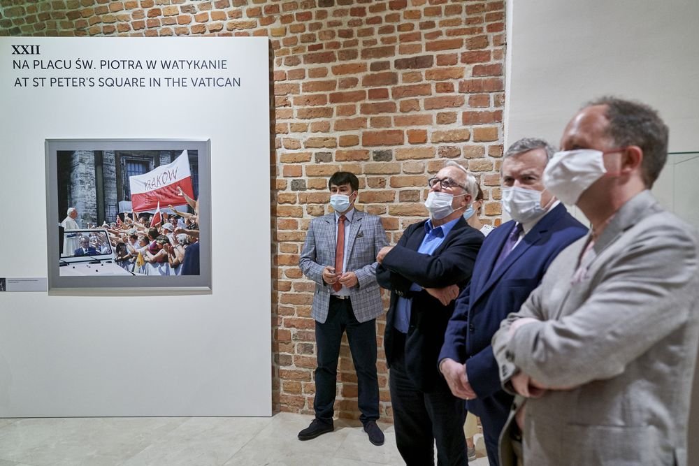 Pięciu mężczyzn w marynarkach i maseczkach stoi przy ceglanej ścianie i ogląda oprawione zdjęcie na białej planszy z napisem "NA PLACU ŚW. PIOTRA W WATYKANIE / AT ST PETER'S SQUARE IN THE VATICAN", na zdjęciu widać tłum z flagą i samochód.