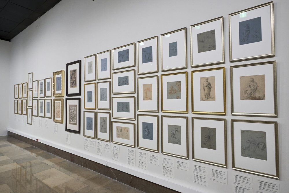 Wnętrze galerii z długą białą ścianą pokrytą równymi rzędami małych oprawionych rysunków i szkiców w złotych, srebrnych i ciemnych ramach, z kartkami informacyjnymi pod obrazami oraz widoczną polerowaną marmurową podłogą i fragmentem sufitu.