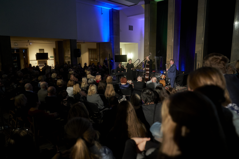 Widok z tyłu na zatłoczoną salę z siedzącą publicznością zwróconą ku małej scenie, gdzie stoją i grają muzycy przy keyboardzie, kontrabasie, zestawie perkusyjnym oraz saksofonie, obok kobieta przy mikrofonie, scena oświetlona niebiesko‑zielonymi reflektorami i wysokimi kolumnami.
