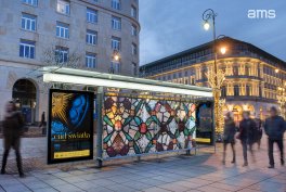 Wieczorny miejski deptak z przystankiem autobusowym o szklanych panelach z kolorowym wzorem przypominającym witraż, podświetlonymi plakatami reklamowymi po bokach, kilkoma rozmytymi przechodniami, brukowanym placem, latarnią i oświetlonymi kamienicami w tle oraz widocznym logo "ams" w prawym górnym rogu.