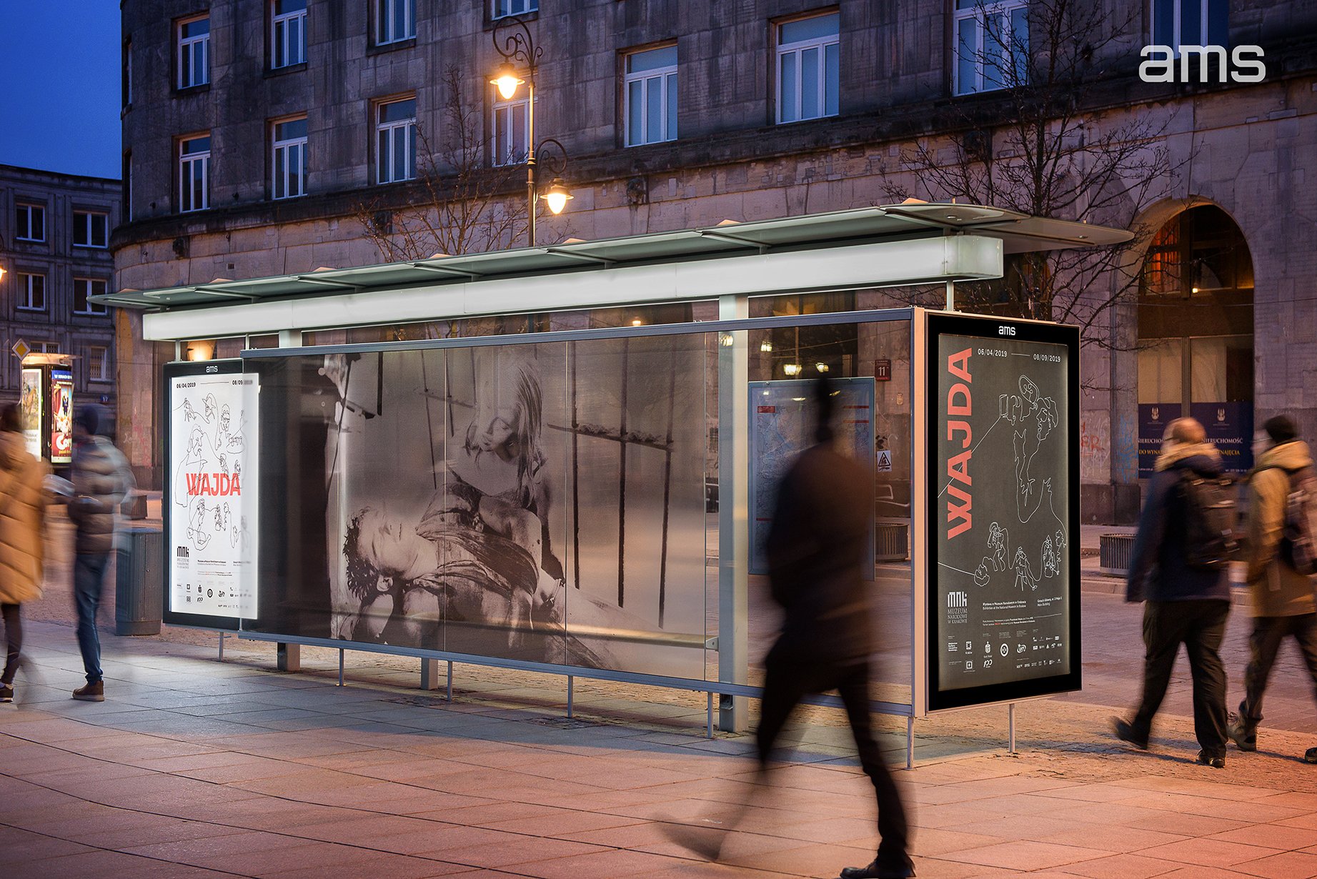 Szklana wiata przystankowa wieczorem na tle kamienicy, z dużą czarno‑białą fotografią na tylnej ścianie, dwiema podświetlonymi pionowymi reklamami z napisem "WAJDA", latarniami ulicznymi i rozmazanymi sylwetkami przechodniów.