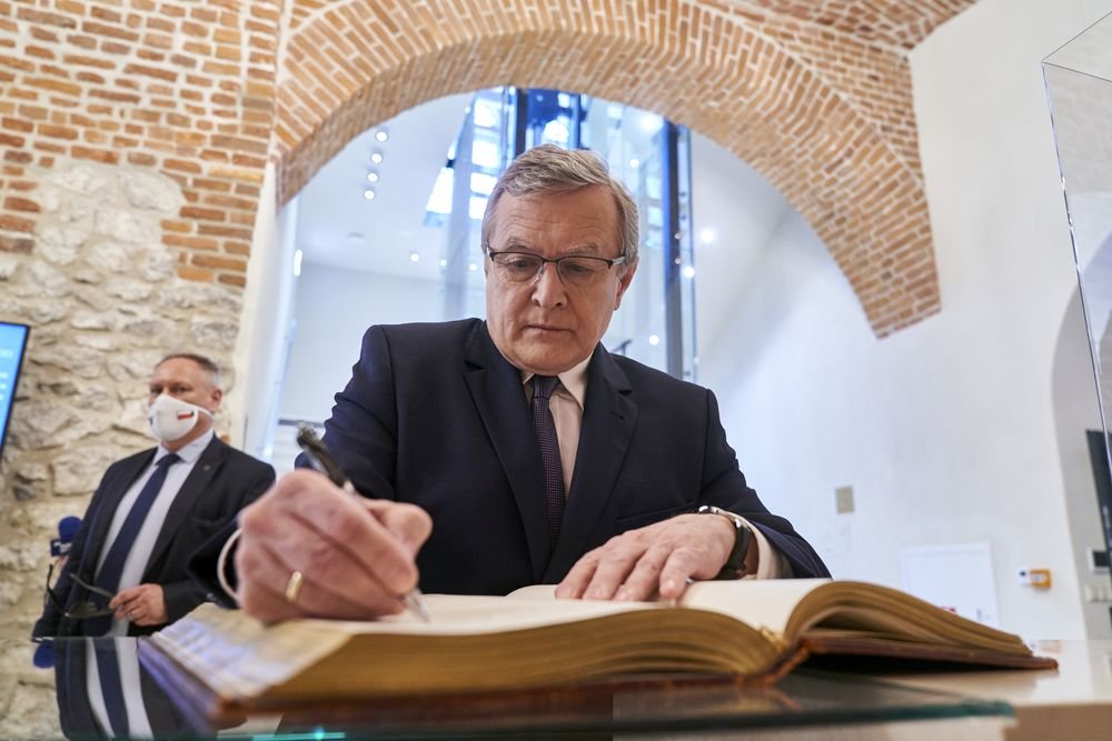 Mężczyzna w garniturze i okularach podpisuje dużą, otwartą księgę leżącą na szklanym blacie w wnętrzu z ceglaną arkadą i kamienną ścianą; po lewej stoi inny mężczyzna w garniturze i maseczce, a w tle widoczna nowoczesna szklana konstrukcja.