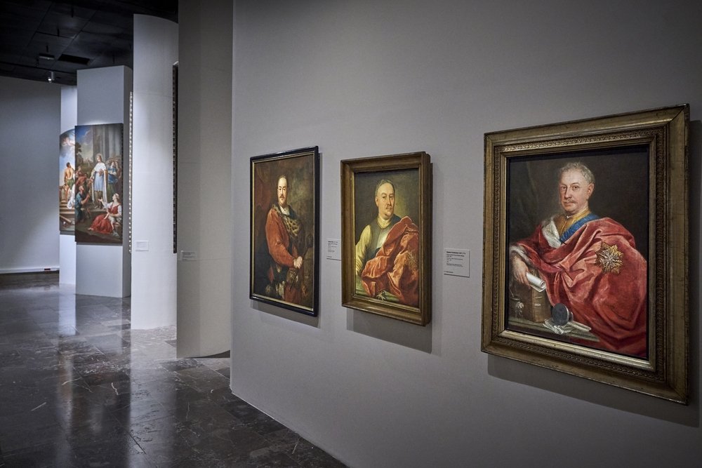Wnętrze galerii sztuki z trzema oprawionymi portretami mężczyzn wiszącymi obok siebie na jasnej ścianie po prawej, po lewej wysokie białe filary z kolejnymi obrazami, ciemna marmurowa podłoga odbijająca światło i punktowe oświetlenie sufitowe.