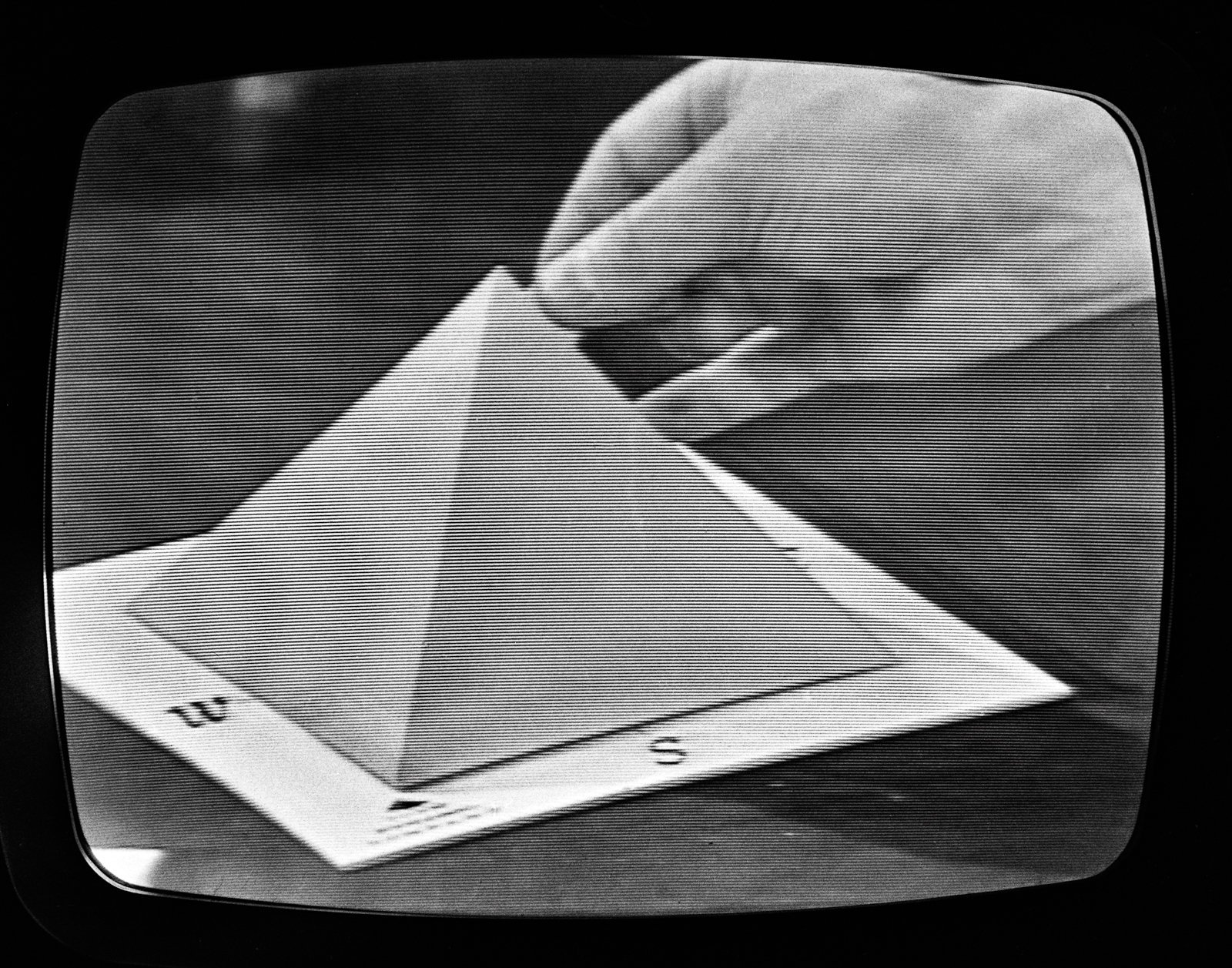 Czarno-białe zdjęcie na ekranie telewizora z poziomymi liniami, pokazujące dłoń układającą papierową piramidę na prostokątnej podstawie z widocznymi literami W i S.