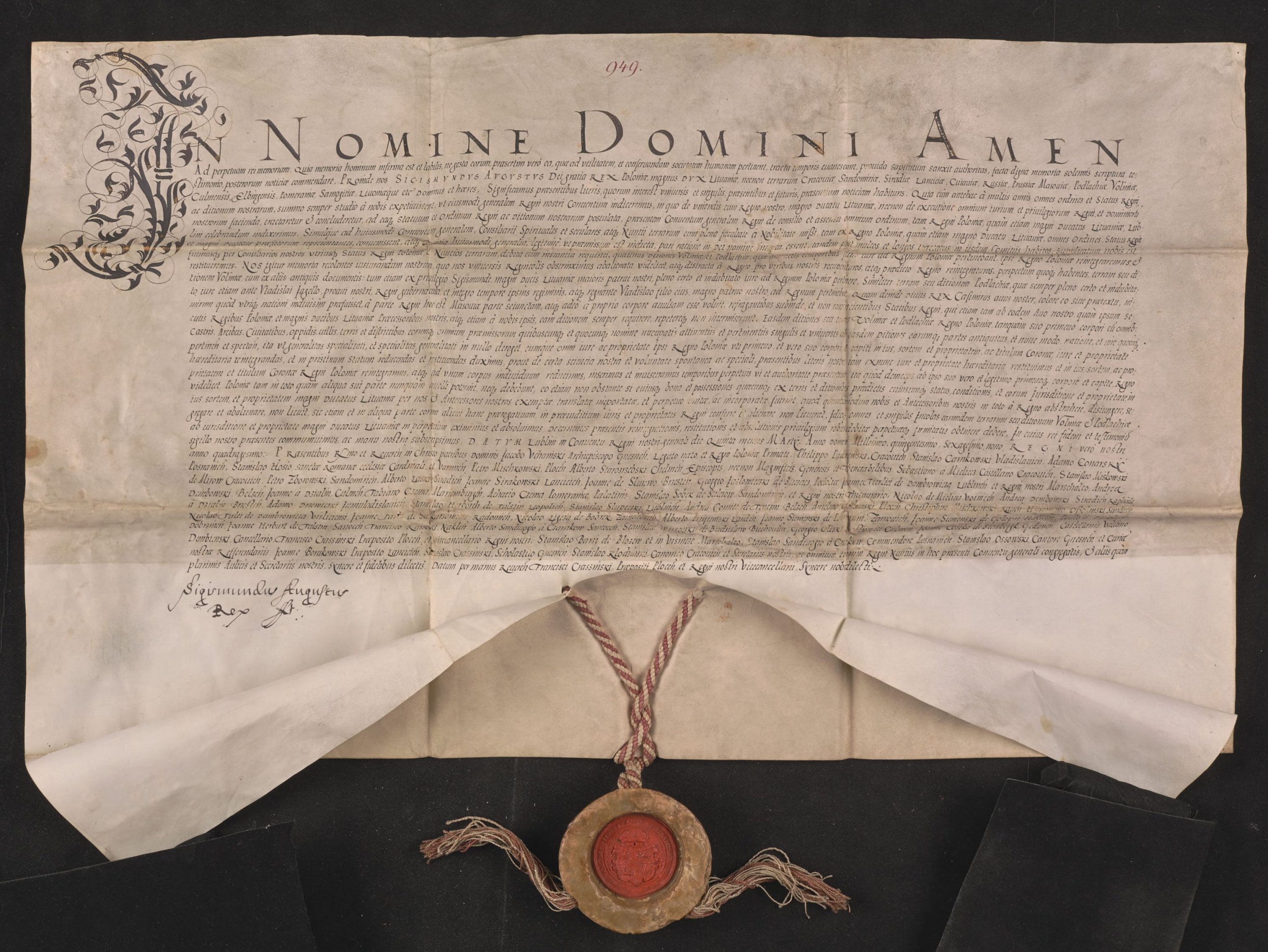 Stary pergaminowy dokument z gęsto zapisanym tekstem i ozdobną inicjałą po lewej, dużym drukowanym nagłówkiem "IN NOMINE DOMINI AMEN" u góry oraz czerwonym numerem "949" nad nim, podpisami w lewym dolnym rogu, złożonymi brzegami i fałdami, na czarnym tle, z centralnie zawieszoną skręconą czerwono-białą sznurką i okrągłą czerwoną pieczęcią woskową osłoniętą pergaminową otoczką na dole.