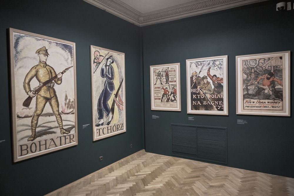 Wnętrze galerii z ciemnozieloną ścianą i pięcioma oprawionymi plakatami historycznymi — od lewej duży plakat żołnierza z karabinem i napisem "BOHATER", obok plakat z postacią i napisem "TCHÓRZ", dalej trzy mniejsze plakaty z ilustracjami i polskimi napisami; widoczna jasna parkietowa podłoga, ozdobna listwa sufitowa, kratka wentylacyjna na ścianie oraz kamera w prawym górnym rogu.