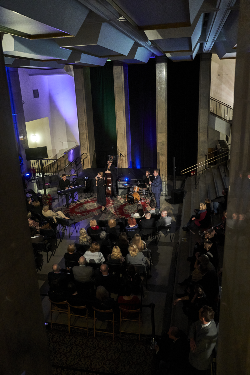Zdjęcie z góry na kameralny koncert w dużej sali z wysokim sufitem i geometrycznymi panelami akustycznymi; scena między trzema wysokimi kolumnami oświetlonymi na niebiesko i fioletowo, na niej keyboardzista, osoba śpiewająca z mikrofonem, kontrabasista, perkusja i stojący muzyk w garniturze, na scenie dywany, przed nią rzędy krzeseł z publicznością, po bokach widoczne schody i balustrady.