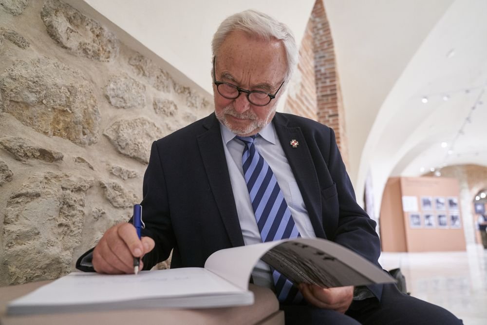 Starszy mężczyzna z siwymi włosami i okularami w ciemnym garniturze i paskowanym krawacie siedzi przy stole w kamiennym wnętrzu, pisze długopem w otwartej księdze trzymając jej strony, w tle sklepione przejście i tablice wystawy.