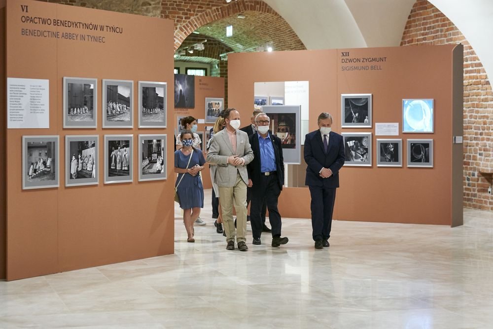 Grupa osób w maseczkach przechodzi przez wystawę w sali z ceglanymi łukowymi sklepieniami, między dużymi terakotowymi panelami z oprawionymi czarno-białymi fotografiami i podpisami (m.in. "Opactwo Benedyktynów w Tyńcu"), na jasnej połyskującej posadzce.
