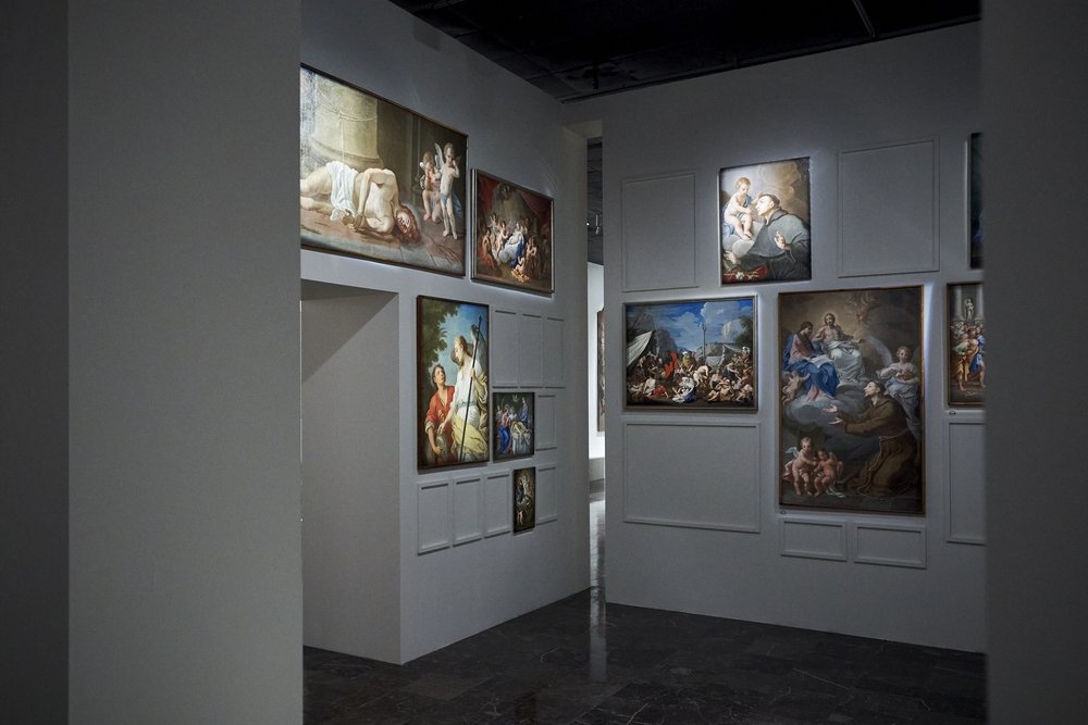 Wnętrze galerii z białymi ścianami i ciemną błyszczącą podłogą, w którym na prawej i środkowej ścianie w panelach powieszono kilkanaście klasycznych obrazów przedstawiających sceny religijne, z przejściem do sąsiedniej sali po lewej i ciemnym sufitem ponad.