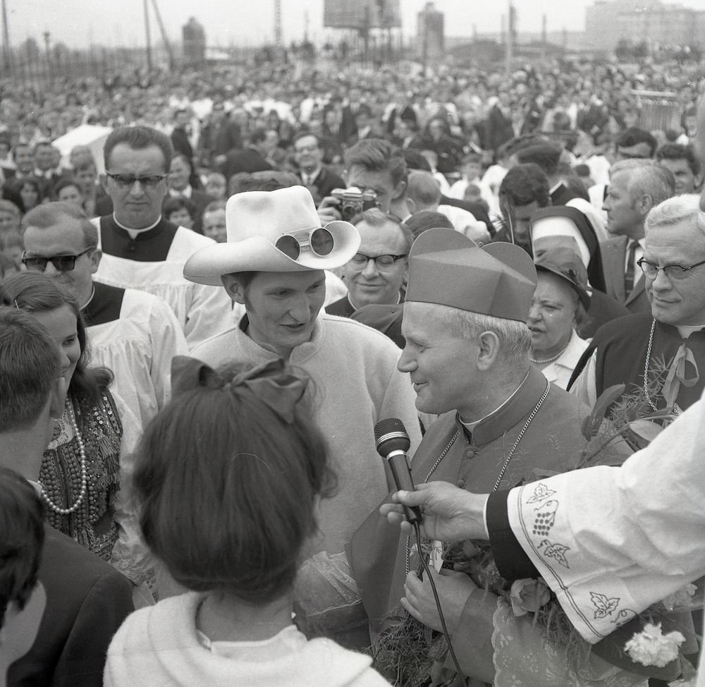 Czarno-białe zdjęcie tłumu na zewnątrz, na pierwszym planie duchowny w nakryciu głowy liturgicznym z mikrofonem rozmawia z mężczyzną w białym kapeluszu z założonymi okularami na rondzie, wokół stoją siostra zakonna, ministranci w albach, osoby w strojach ludowych i inni zgromadzeni, a w tle rozległy tłum.