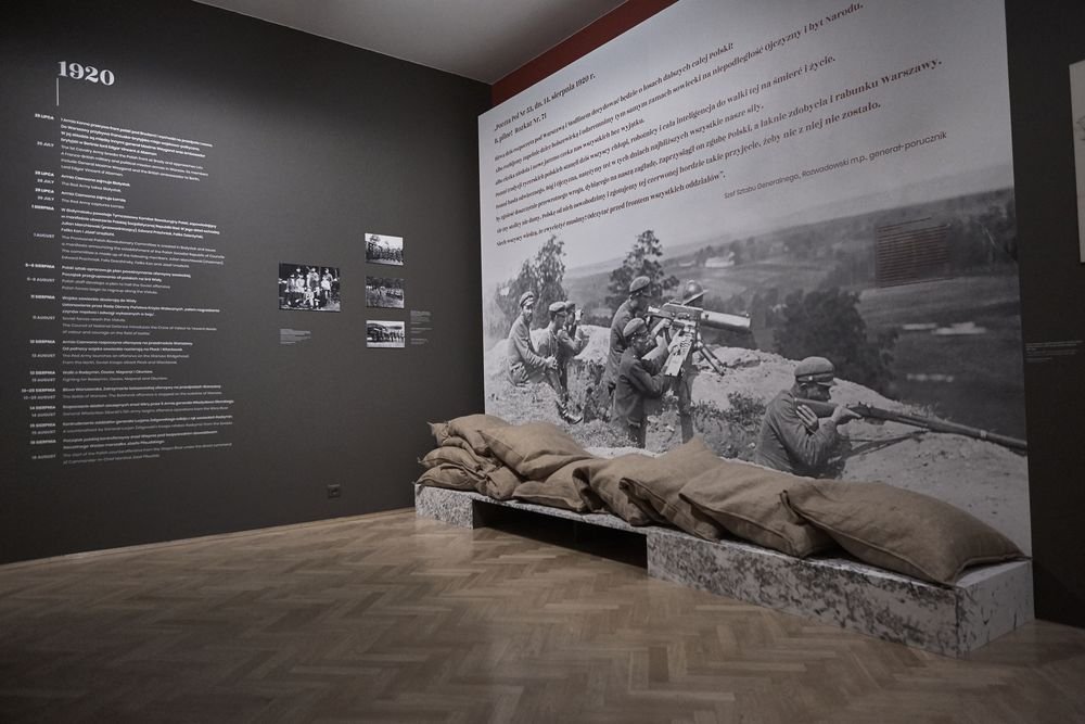 Wnętrze wystawy muzealnej z dużym napisem „1920” na ciemnej ścianie ze szczegółową linią czasu i małymi fotografiami po lewej oraz wielkoformatową czarno-białą fotografią żołnierzy w okopie na prawej ścianie, przed którą ułożono stos worków z piaskiem na niskim podwyższeniu na jasnej drewnianej podłodze.