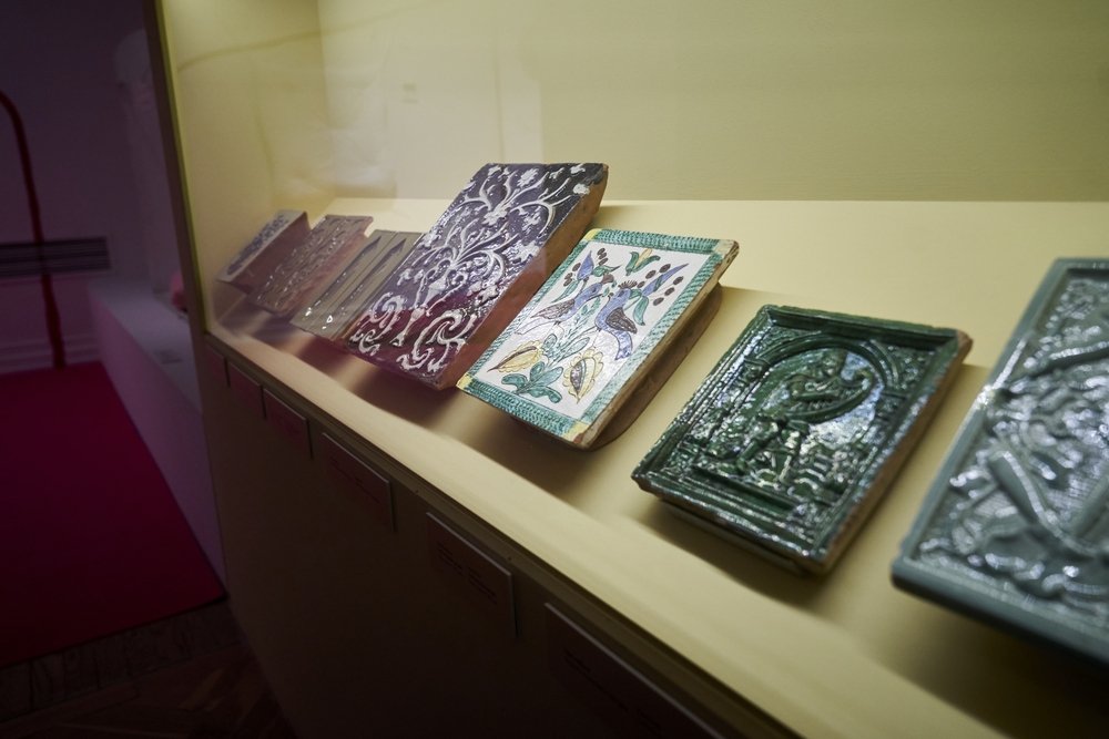 Rząd ozdobnych, glazurowanych płytek ceramicznych o różnych wzorach (zielona płytka z motywem figury w arkadzie, kolorowa z ptakami i liśćmi, większa ciemna z białymi ornamentami) ustawionych pochyło w szklanej gablocie na żółtym tle ekspozycji.