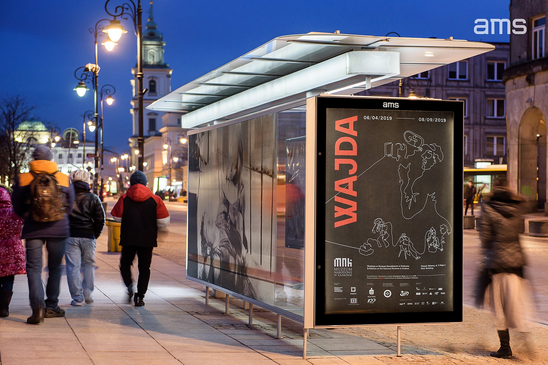 Przystanek z przeszkloną wiatą i podświetloną gablotą reklamową z dużym pionowym napisem "WAJDA", stojący wieczorem przy oświetlonej ulicy z kilkoma przechodniami na chodniku oraz zabudową miejską i wieżą kościelną w tle.