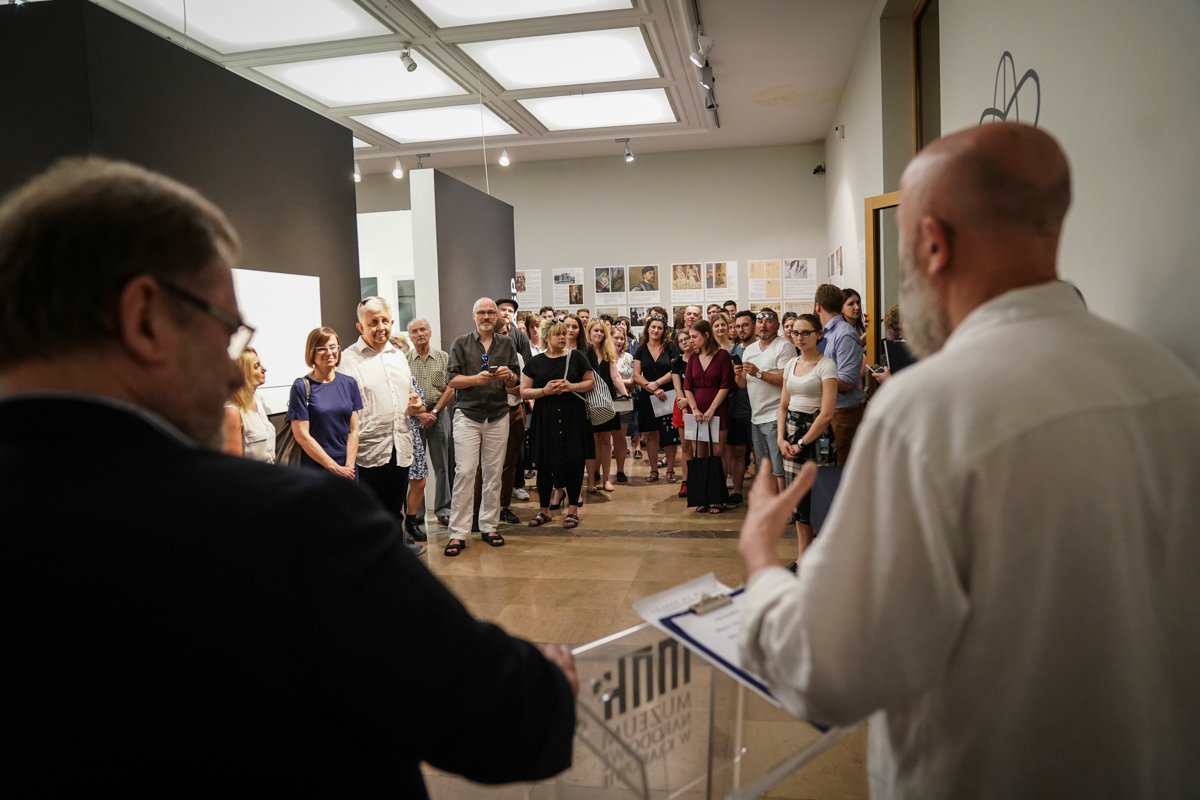 Sala wystawowa z jasnym sufitem panelowym, tłumem osób w strojach codziennych ustawionych naprzeciw dwóch mężczyzn na pierwszym planie przy przezroczystej mównicy — jeden z mężczyzn gestykuluje i trzyma dokumenty, na ścianach w tle widoczne plakaty i prace wystawowe.
