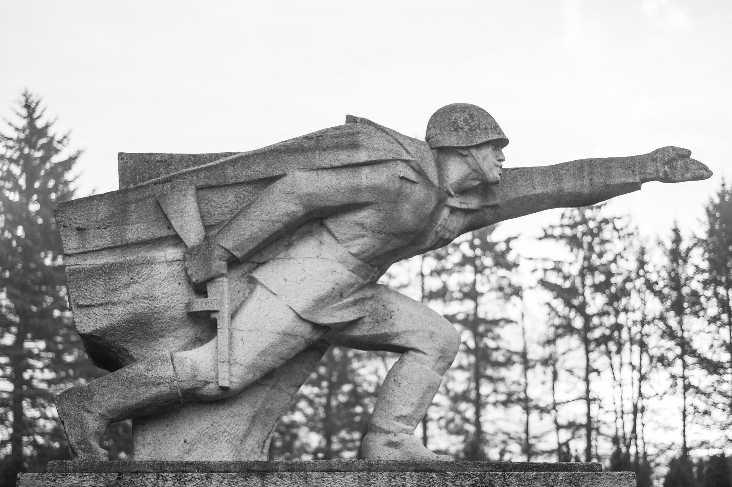Czarno-białe zdjęcie kamiennego pomnika przedstawiającego postać w hełmie i pelerynie, pochyloną do przodu z wyciągniętą prawą ręką i trzymanym na plecach karabinem, osadzonego na kamiennej podstawie na tle drzew.