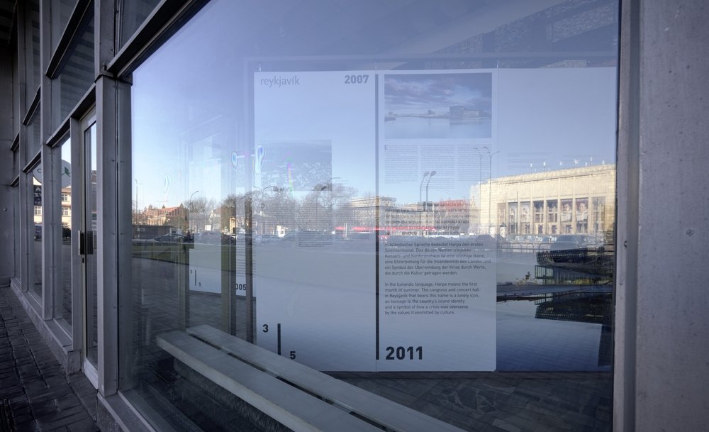 Duże szklane okno budynku odbijające miejski widok: w szybie widoczna wewnętrzna plansza z tekstem i datami „2007” i „2011” oraz ławka, a w odbiciu plac z ulicą i okazały budynek z kolumnami, po lewej ciąg betonowych słupów i chodnik.