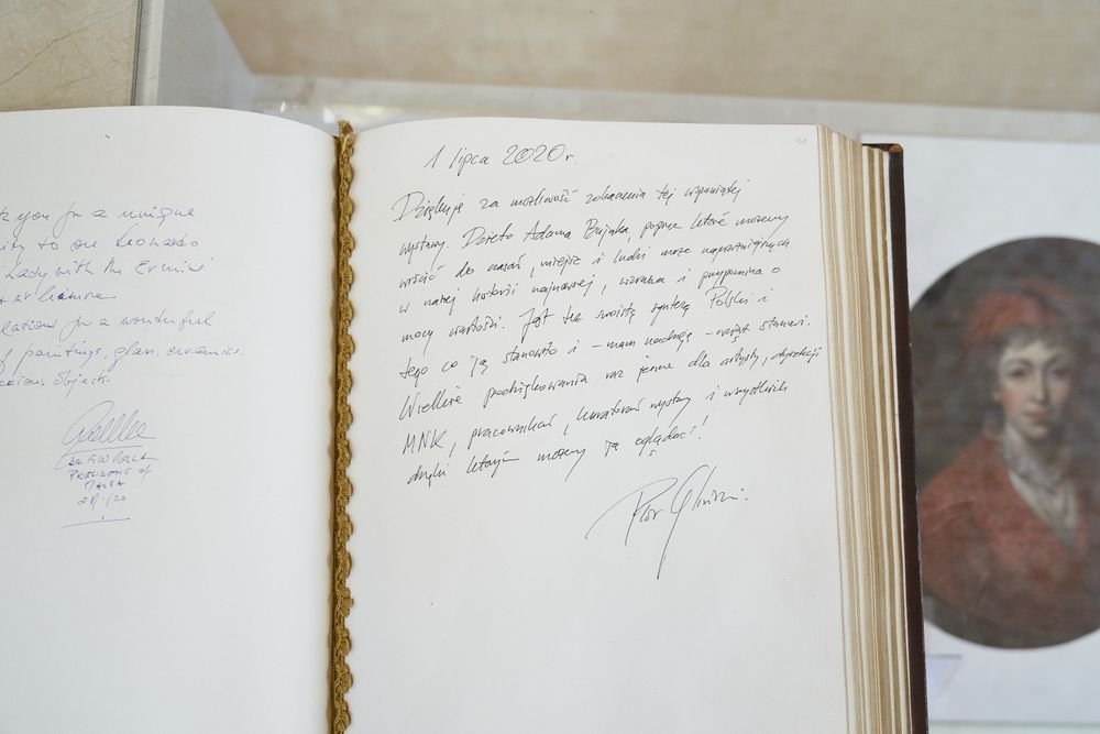 Otwarta duża książka z odręcznymi wpisami po polsku datowanymi "1 lipca 2020", złotą plecioną zakładką między stronami, widocznym fragmentem innego wpisu po lewej i rozmytym okrągłym portretem osoby w czerwonym ubraniu po prawej.